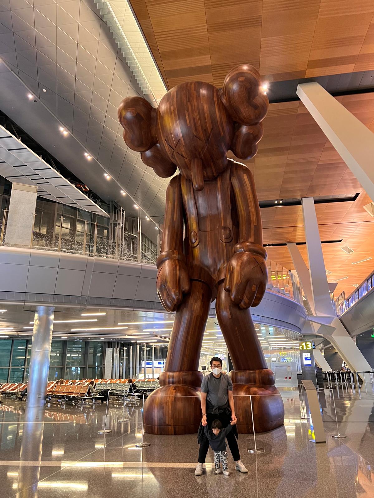 A man and boy taking a photo in front of the Small Lies sculpture by KAWS at the Hamad International Airport, Doha